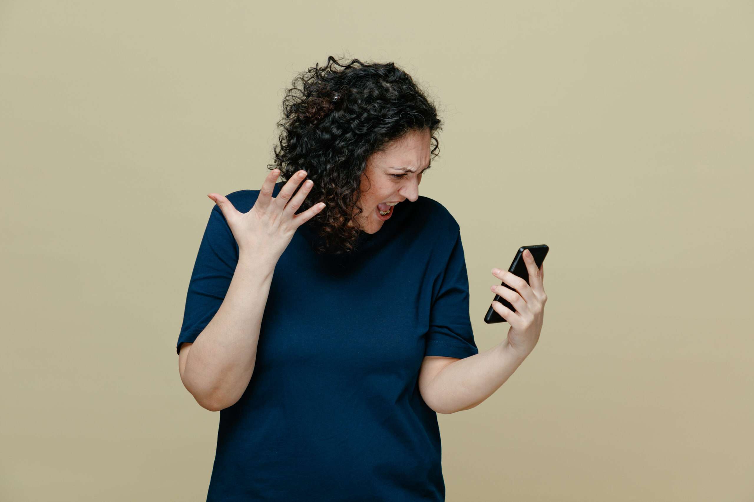 Angry middleaged woman wearing tshirt holding mobile phone looking at it showing empty hand shouting isolated on olive g