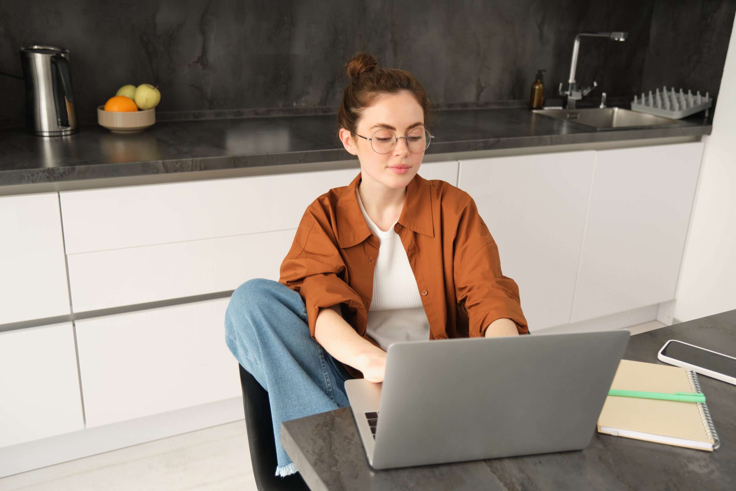 Portrait of young woman student in kitchen sitting with laptop working from home studying or doing