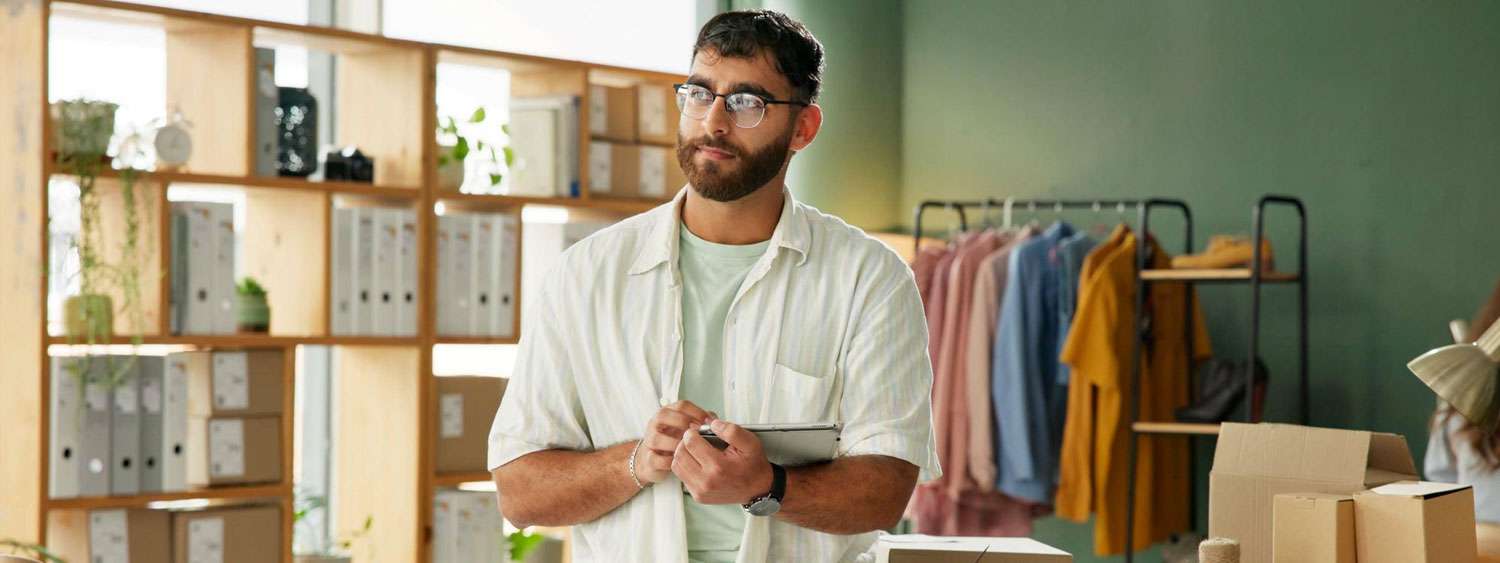Shop owner in glasses holding a tablet in his clothing store, shelves and boxes behind him